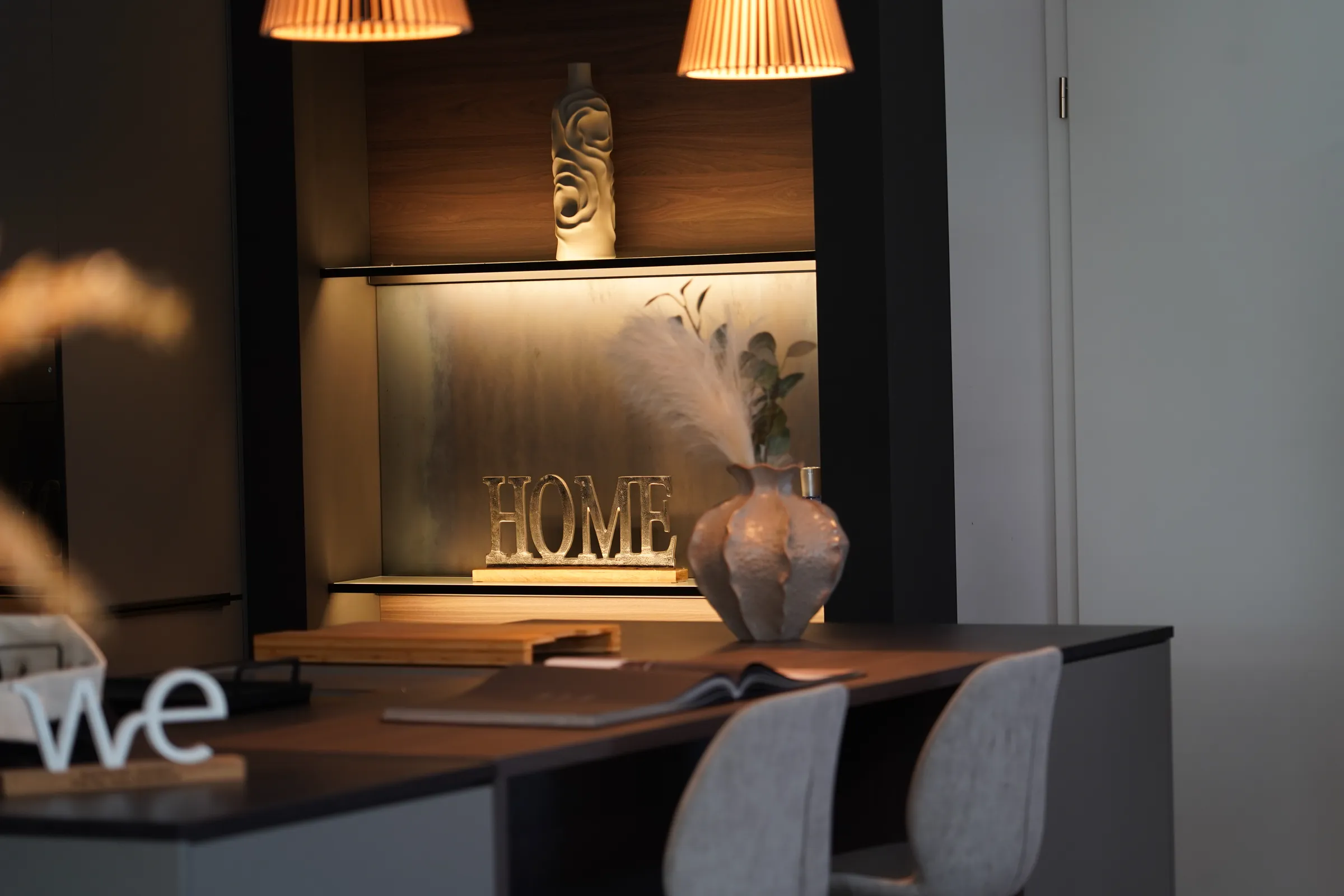 Beleuchtete Wandnische mit dem Schriftzug HOME über einem Tisch mit Stühlen in der Ausstellung
